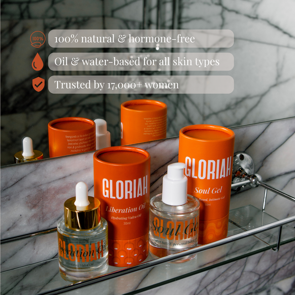 GLORIAH Soul Gel and Liberation Oil products arranged on a glass bathroom shelf with a marble background. The set includes orange cylindrical tubes labeled GLORIAH and a clear bottle with a gold cap labeled GLORIAH