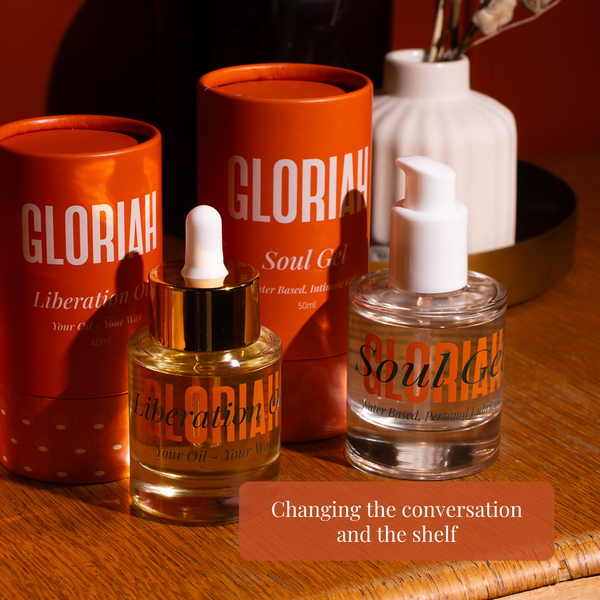 GLORIAH Liberation Oil & Soul Gel products arranged on a wooden surface: two orange cylindrical tubes and a clear glass bottle with a white pump and a smaller bottle with a gold dropper cap. The products are branded GLORIAH, with text including 'Soul Gel' and 'Liberation Oil.