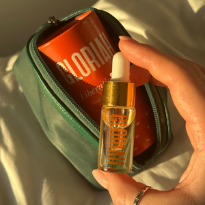 A hand holds a small glass dropper bottle with a gold cap and white dropper over an open mint green zip pouch containing an orange bottle labeled GLORIAH