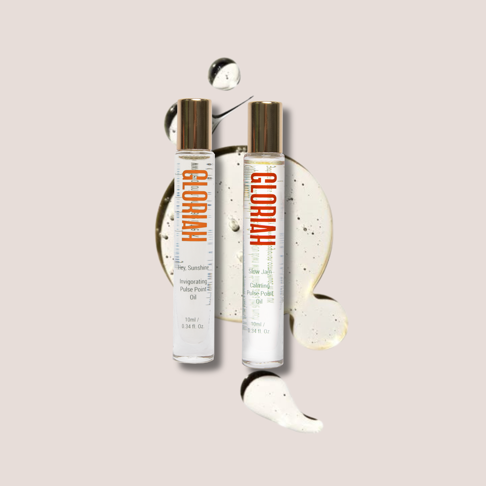 Two clear cosmetic bottles with metallic rose-gold caps and orange 'GLORIAH' branding, shown on a light beige background with decorative cream swirls and a small droplet.