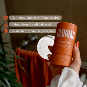 A hand holds a cylindrical orange bottle labeled 'GLORIAH Soul Gel' in front of a bathroom-like background with a towel and plant. White text badges on the left highlight product benefits: hormone, paraben & fragrance free; natural formula, pH balanced; plastic free, premium packaging.