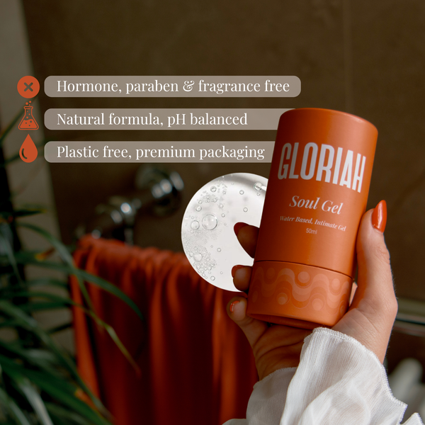 A hand holds a cylindrical orange bottle labeled 'GLORIAH Soul Gel' in front of a bathroom-like background with a towel and plant. White text badges on the left highlight product benefits: hormone, paraben & fragrance free; natural formula, pH balanced; plastic free, premium packaging.