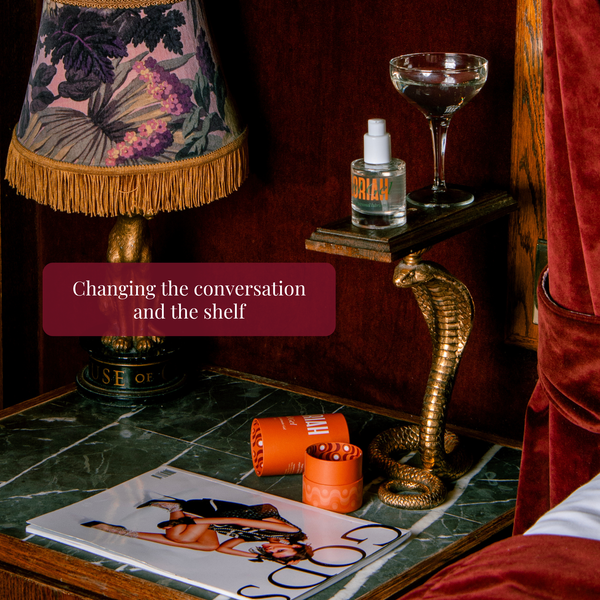 A styled interior scene showing a brass cobra-shaped side table on a green marble nightstand, holding a clear coupe glass and a small bottle labeled 'Soul Gel'. A lamp with a floral patterned shade, a magazine, and orange cylindrical objects are also visible.